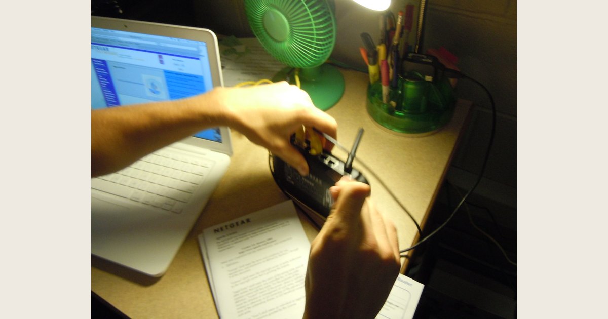 A person connects cables to a black router on a desk beside a laptop.